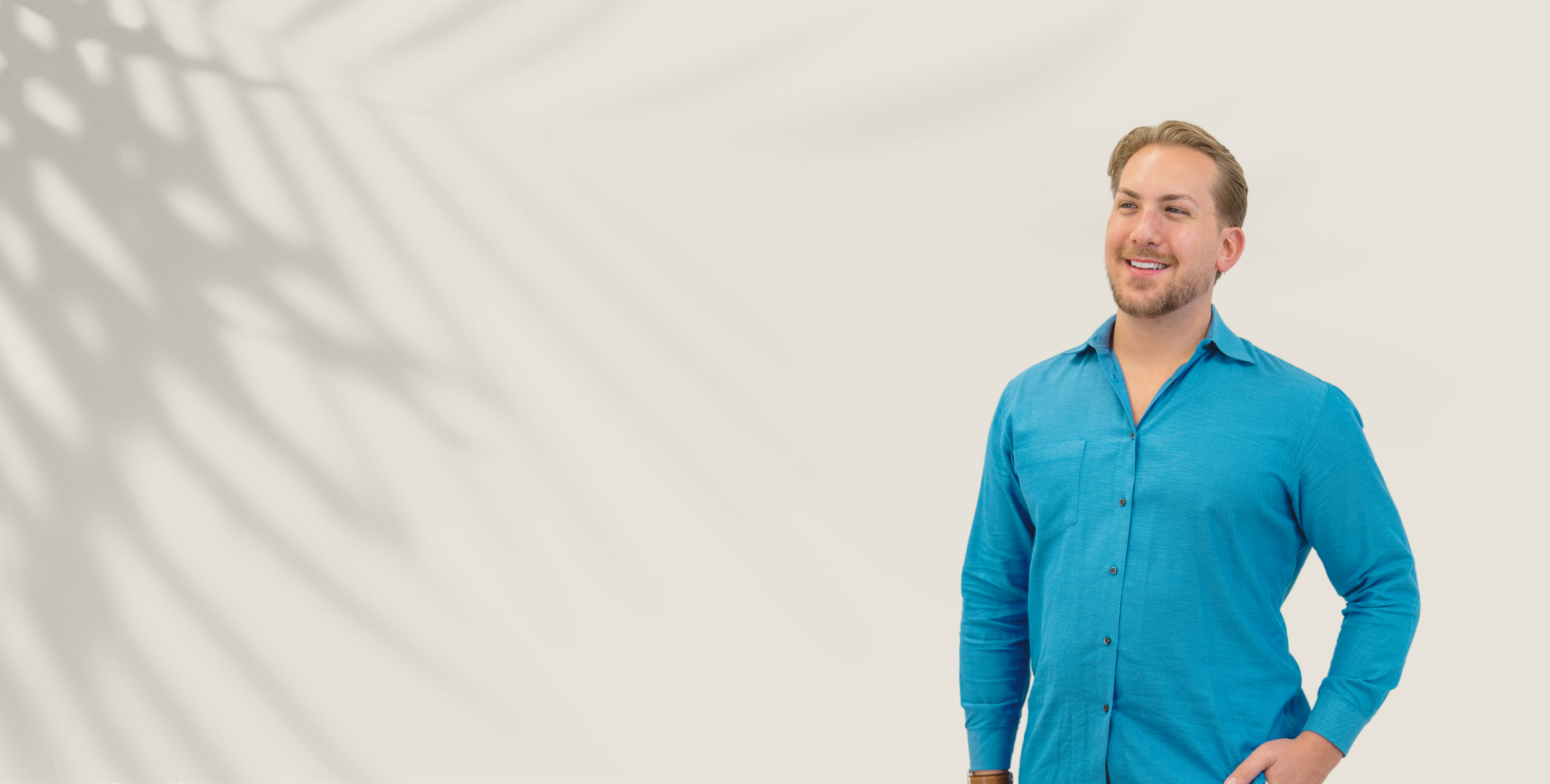 A smiling man with blond hair and a beard in a blue button-down shirt, with palm-leaf shadows on the wall behind him.