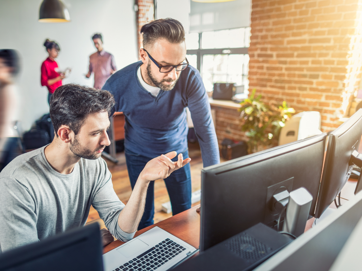 Two men collaborate at computers in a modern office, others blurred in the background.