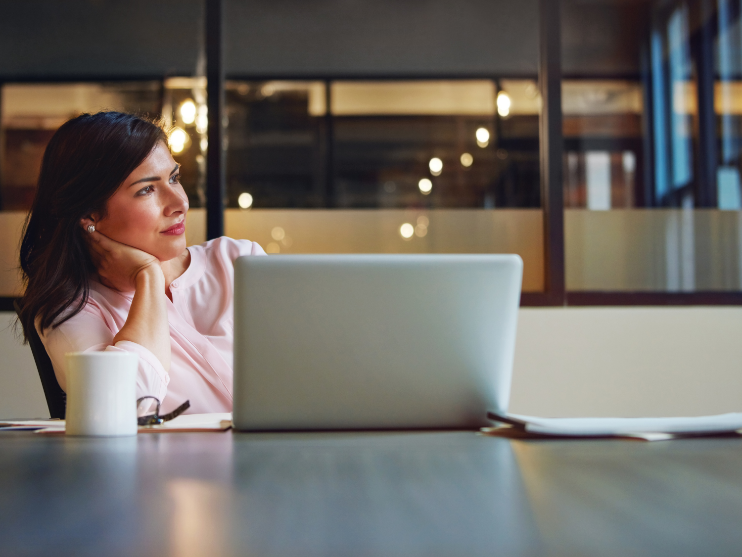 A woman in a pink blouse rests her chin on her hand, looking away from her laptop.