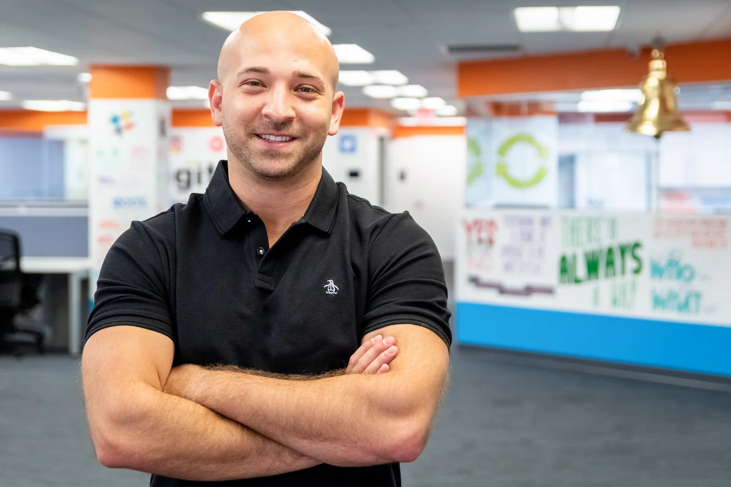 Bald man in a black polo shirt with arms crossed, smiling in an office with motivational posters on the wall.