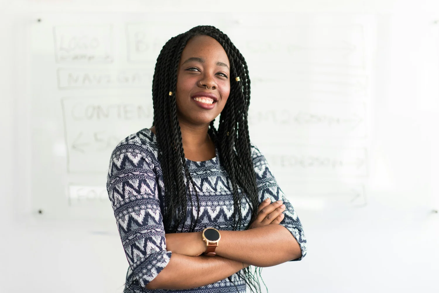 A Black woman with braided hair smiles at the camera with her arms crossed, wearing a patterned top and a watch.