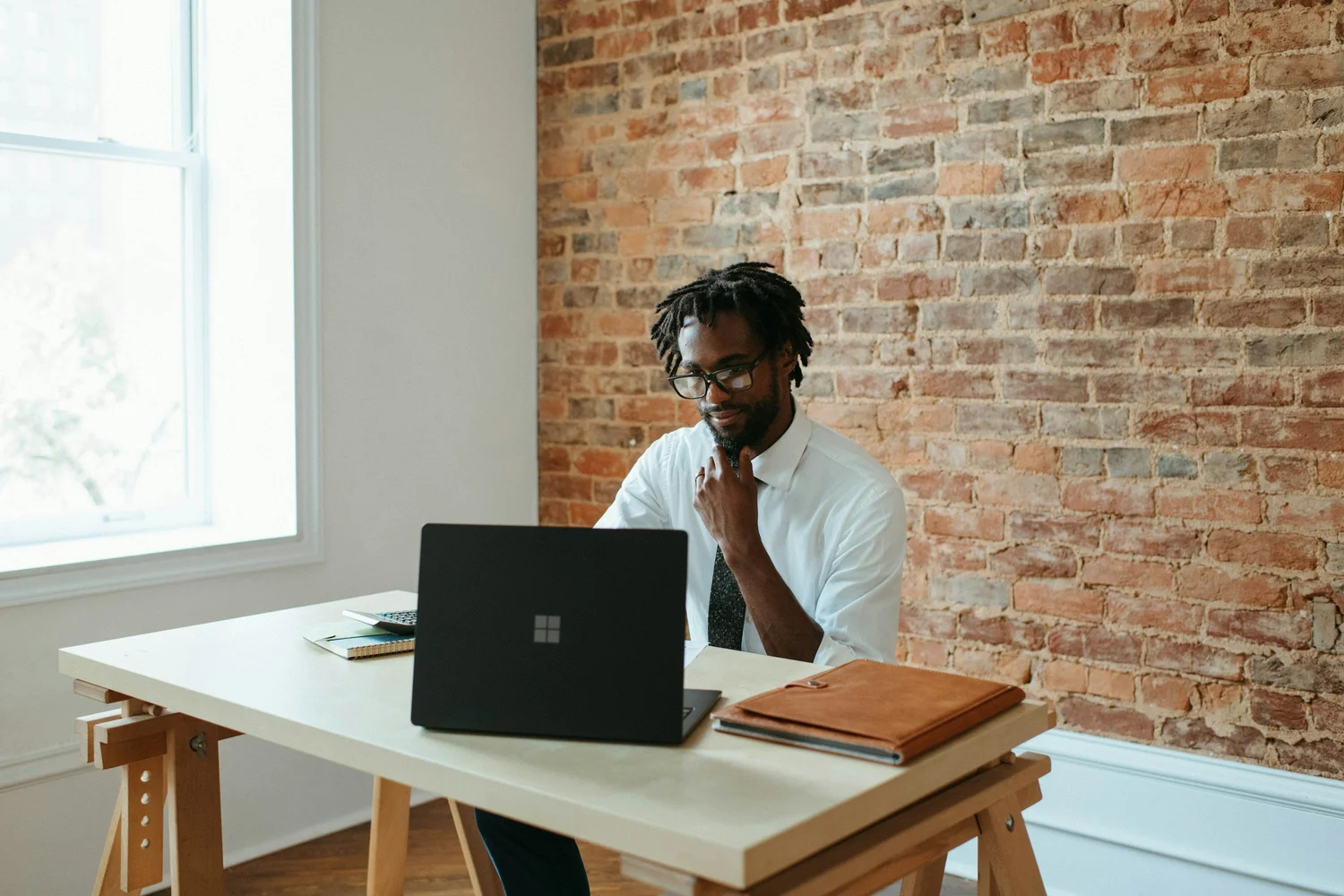 Man with dreadlocks and glasses in a white shirt and tie works on a laptop at a desk.