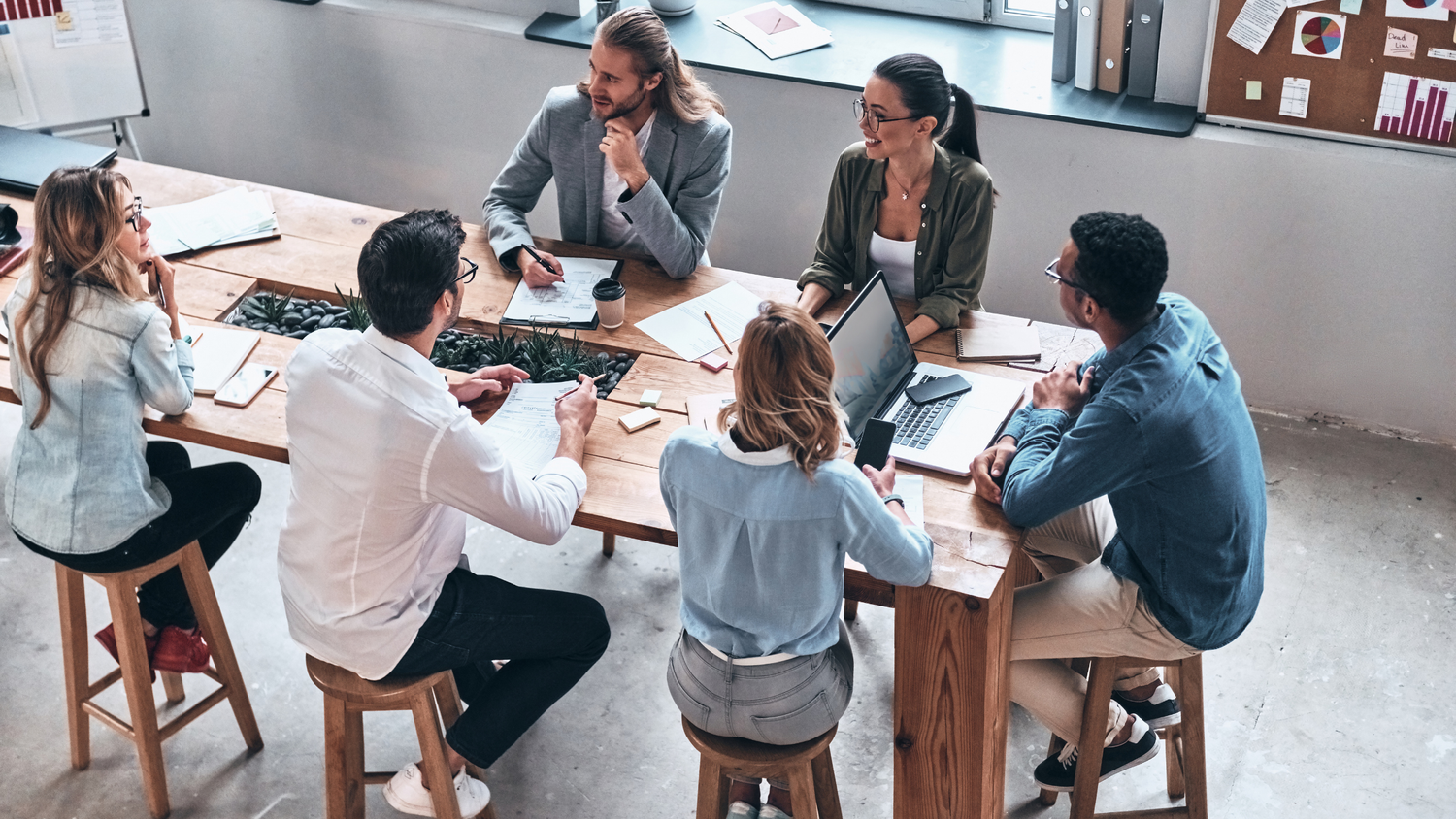 Six people sit around a modern wooden conference table, engaged in discussion with laptops and papers.