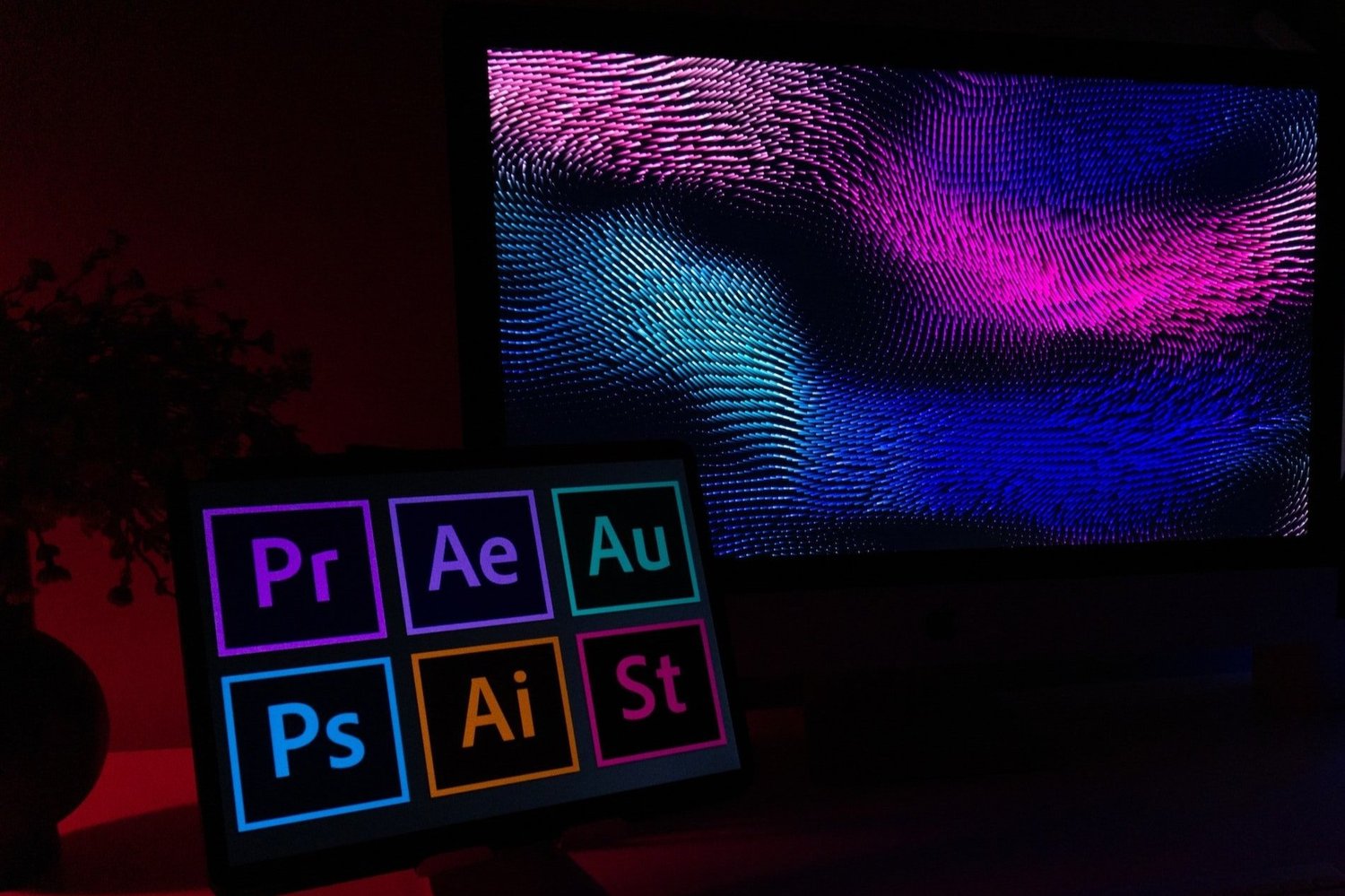 A tablet displays colorful icons for Adobe Premiere, After Effects, Audition, Photoshop, and Illustrator; a large monitor behind it shows a dynamic, colorful design.