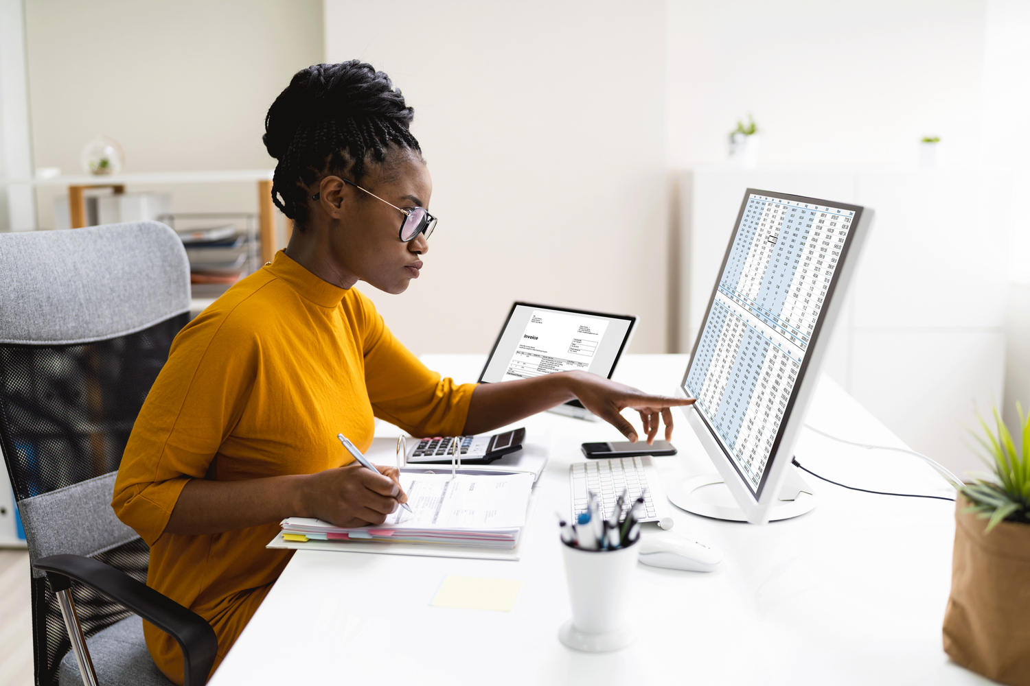 Woman in a yellow shirt and glasses points to a spreadsheet on a monitor while writing on paper, a laptop open nearby.