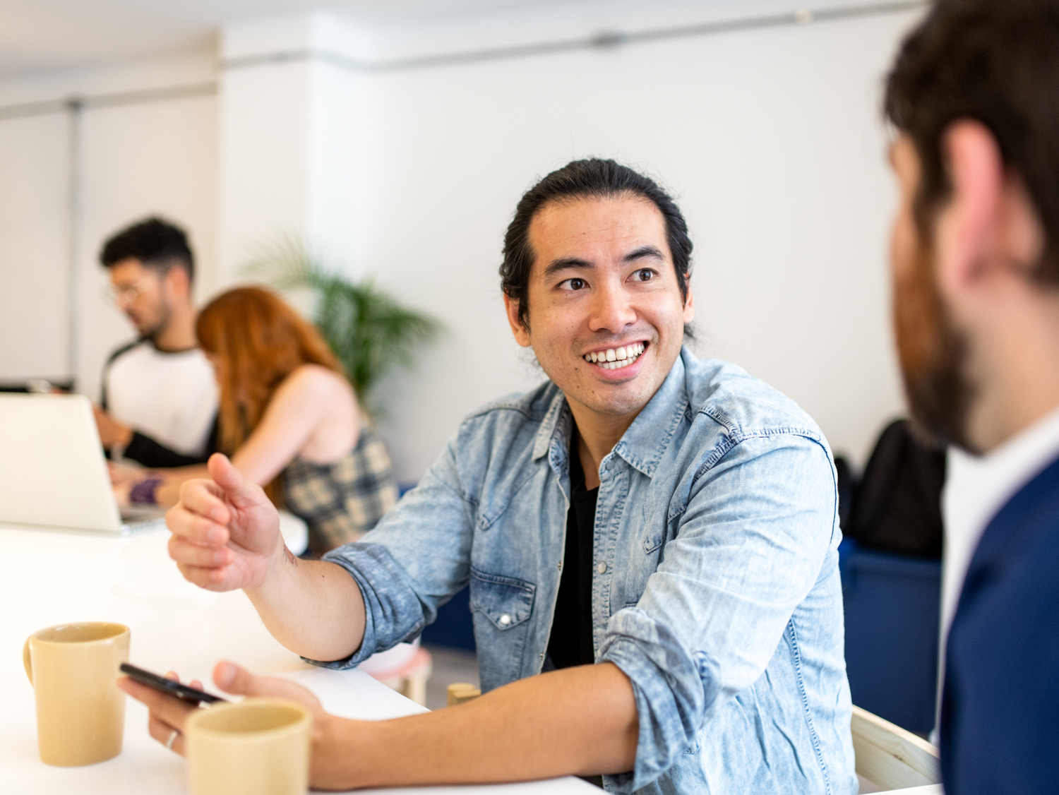 An Asian man in a denim shirt smiles and gestures while talking to someone off-camera in an office, two mugs on the desk.