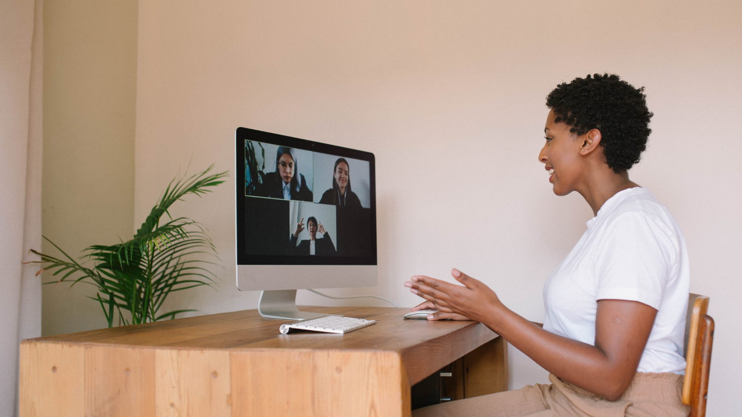 A Black woman in a white t-shirt smiles and gestures while on a video call with three colleagues on her screen.