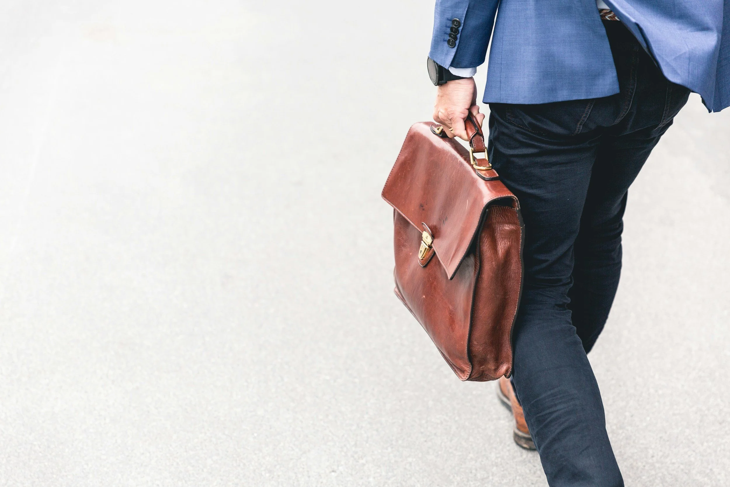 Close-up of a businessman in a blue suit jacket and black pants carrying a brown leather briefcase and wearing a smartwatch.
