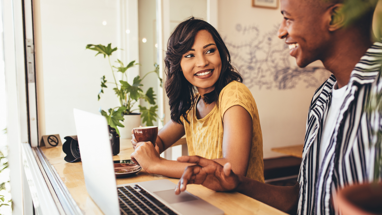 A Black woman in a yellow shirt smiles holding a coffee cup and looking at a laptop while a Black man in a striped shirt gestures toward it.