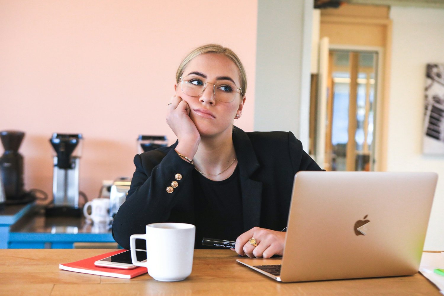 A businesswoman with glasses rests her chin on her hand, looking bored while working on a laptop in an office.