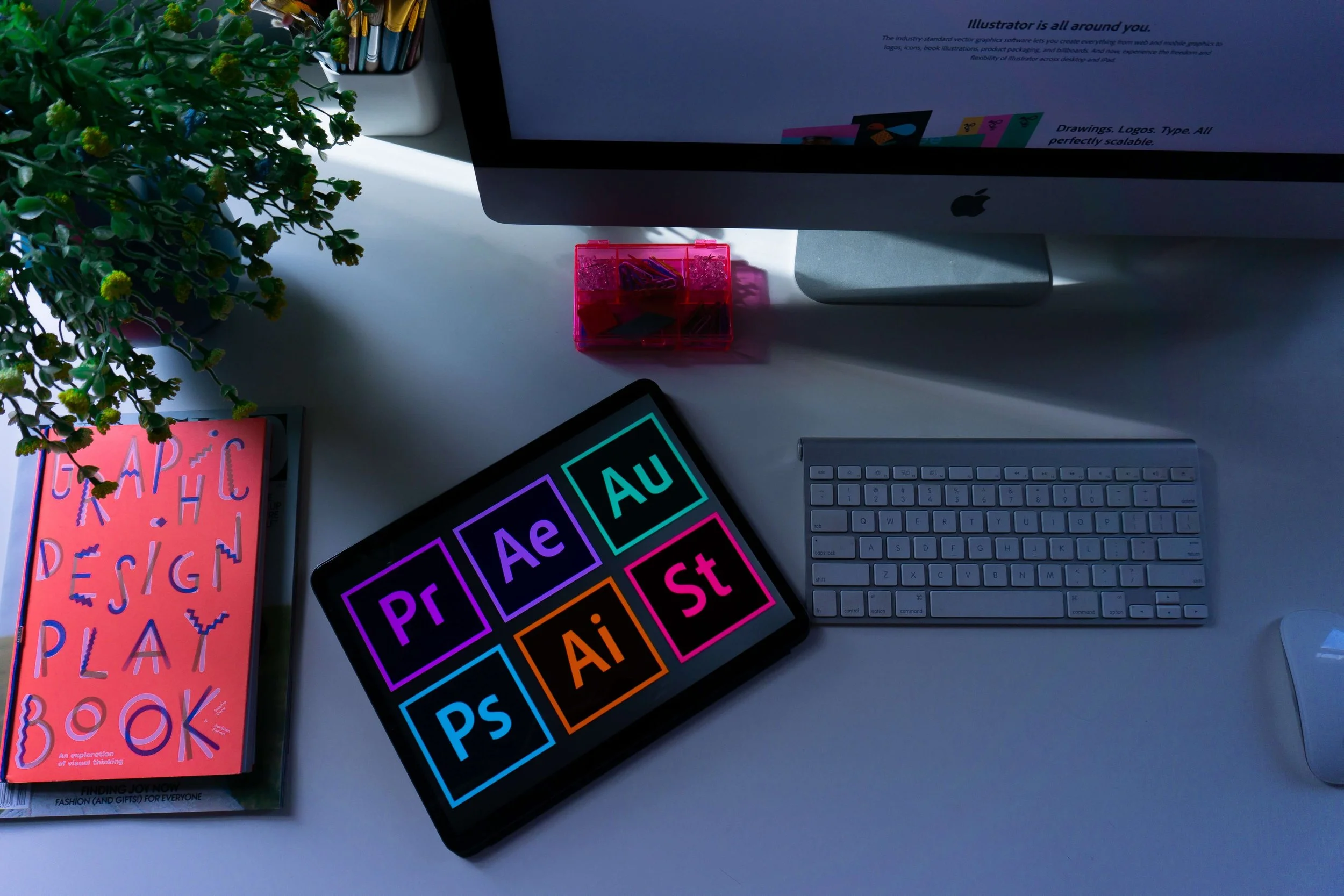 Tablet displaying Adobe Creative Cloud icons including Premiere Pro, After Effects, Audition, Photoshop, Illustrator, and Stock, beside a graphic design book and keyboard.