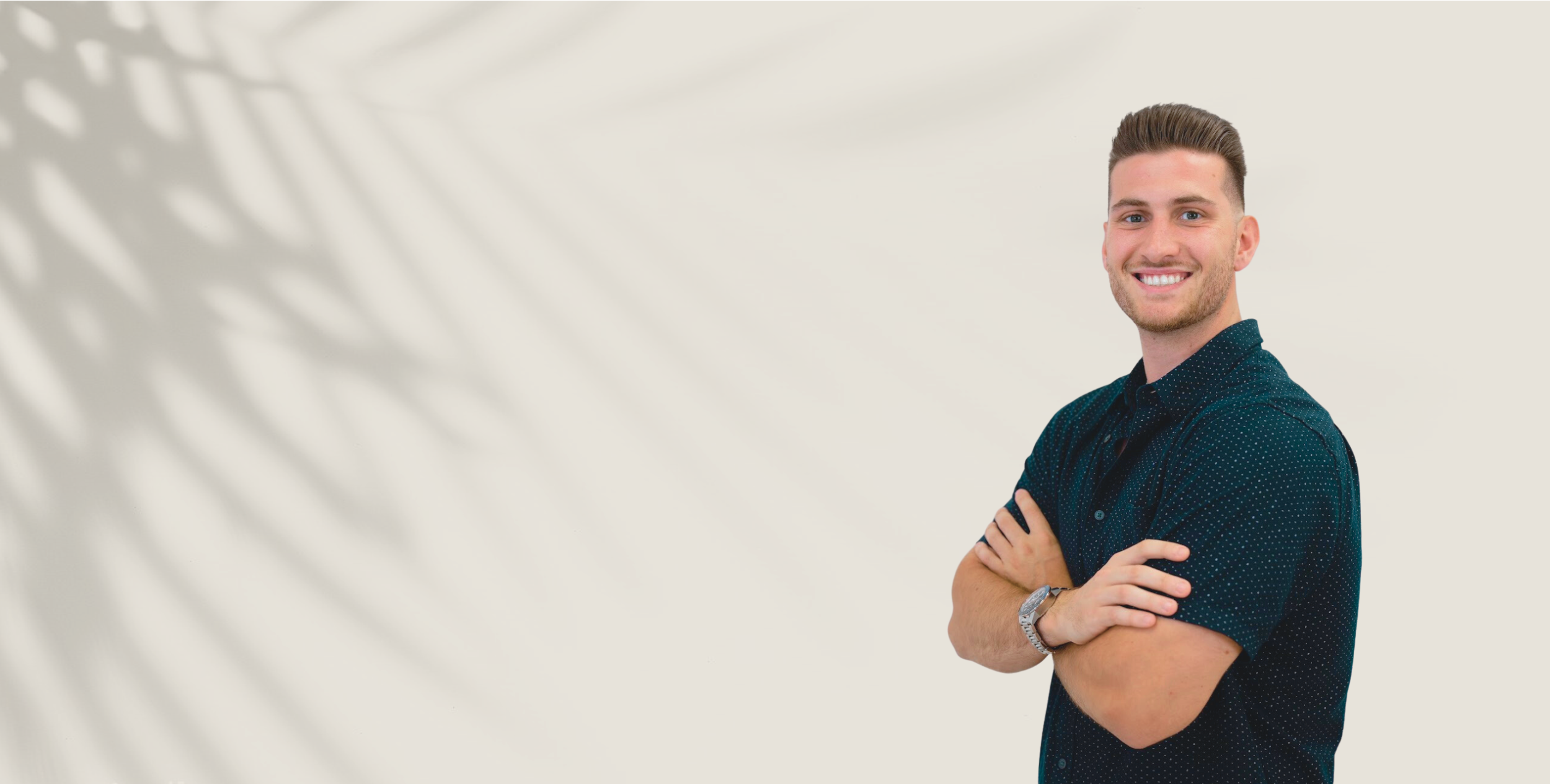 A smiling man with short brown hair in a dark blue patterned short-sleeved shirt and a watch, standing with his arms crossed.