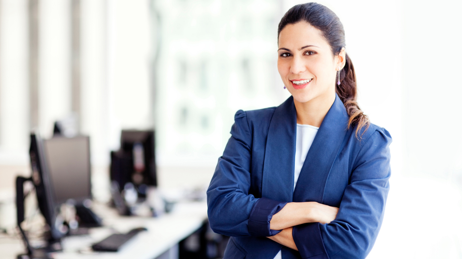 Smiling businesswoman with arms crossed in an office, her dark hair pulled back in a ponytail.