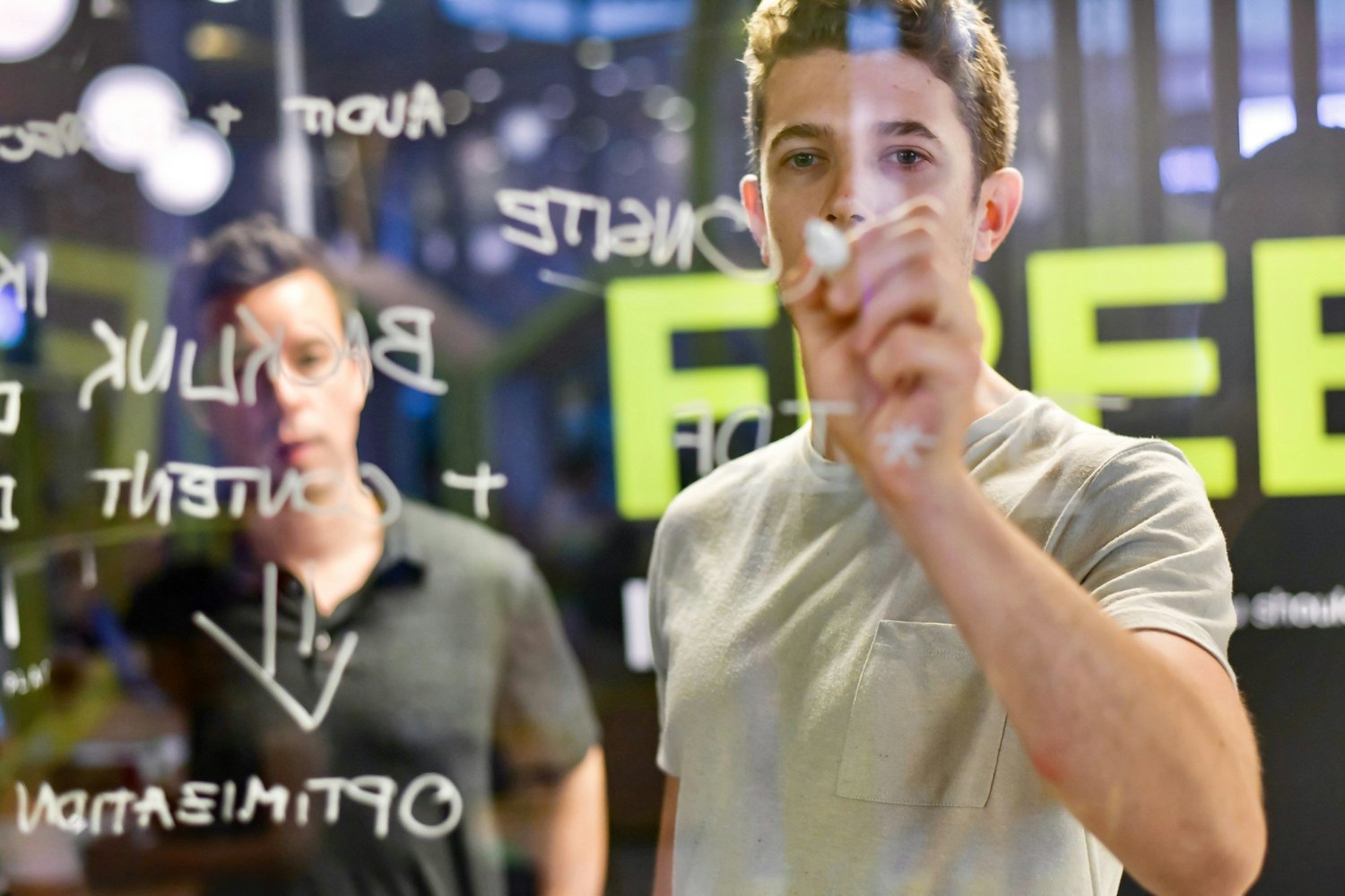 A young man writes on a glass board as another man watches, with the word FREE visible in bright green letters.