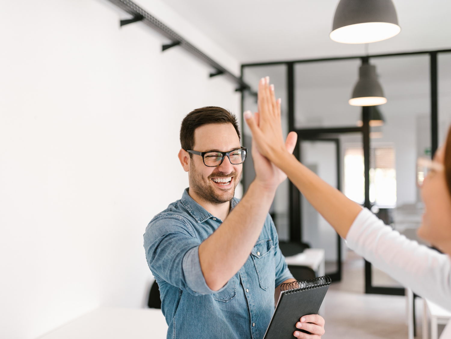 Man in glasses and a denim shirt happily gives a coworker a high five while holding a notepad.
