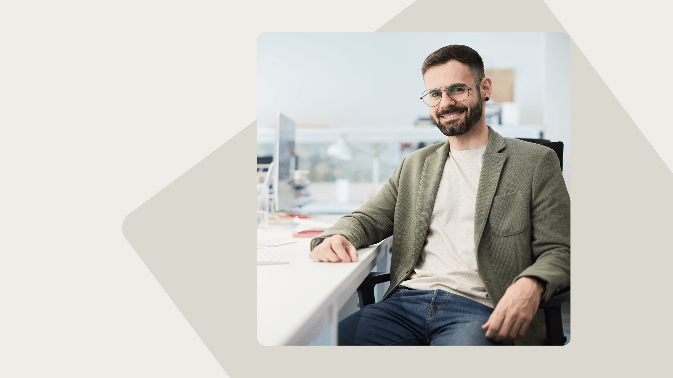 Man with glasses and a beard wearing a green jacket and jeans, smiling while sitting at a desk in an office.