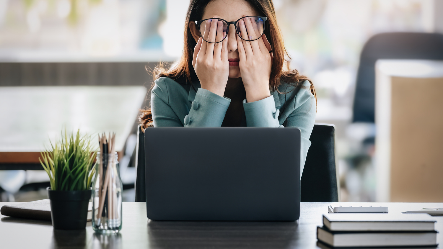 Woman wearing glasses rubs her eyes, sitting at a desk with a laptop and plant, looking tired.