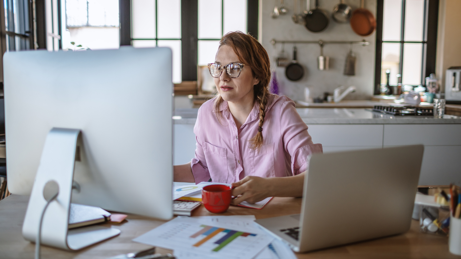 Woman with glasses and braided hair working at a desk with a desktop computer, laptop, and red mug.