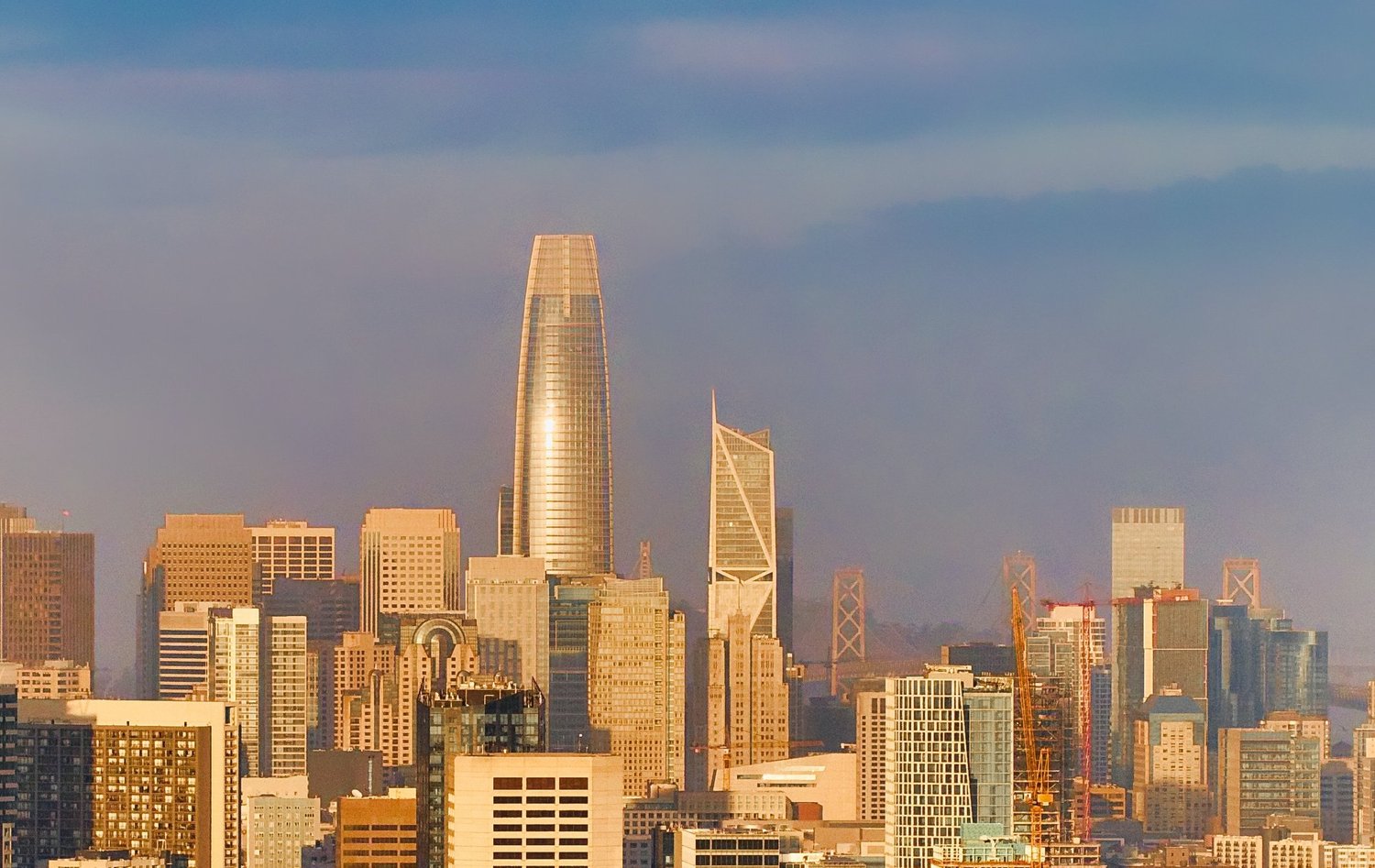 The San Francisco skyline at sunset, featuring the Salesforce Tower and other skyscrapers, with a bridge in the distance.