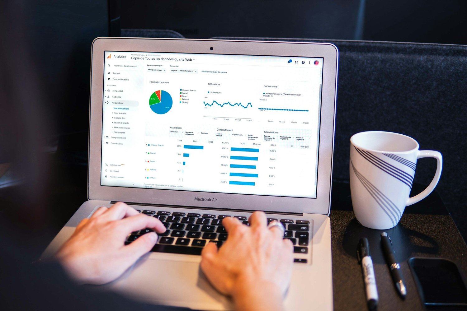 Person typing on a MacBook Air displaying Google Analytics data showing traffic sources, user behavior, and conversion rates.