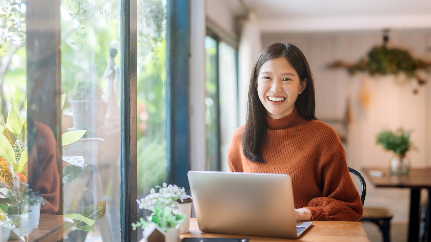 A smiling young Asian woman in an orange sweater sits at a cafe table with a laptop.
