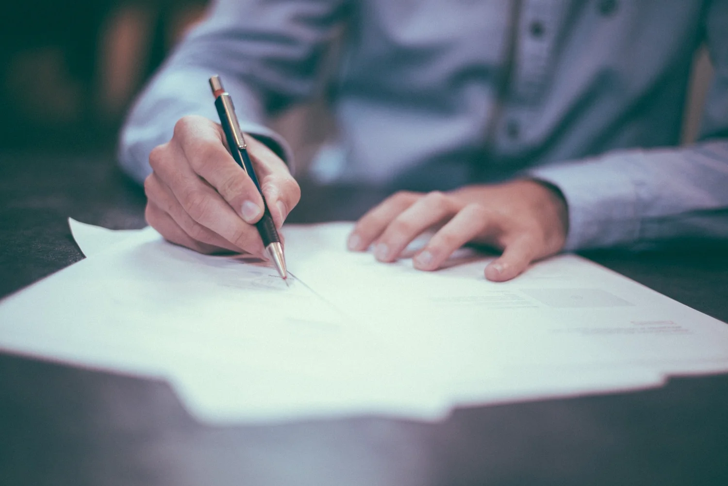 Close-up of a person in a blue shirt writing on a white sheet of paper with a pen.