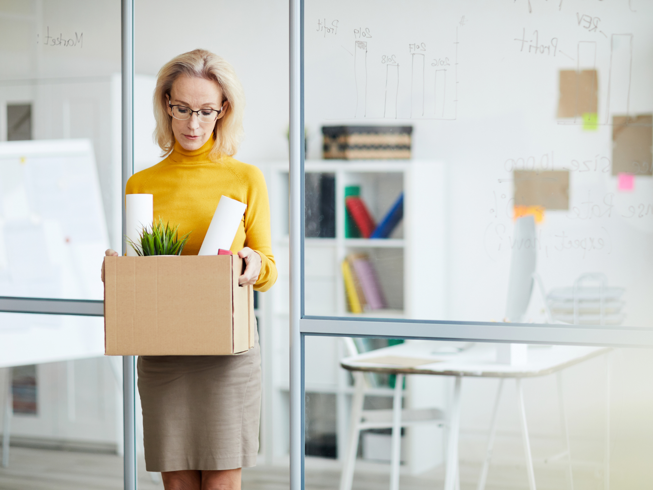 Woman in a yellow sweater holding a box of office supplies, including a plant and rolled papers, looking down.