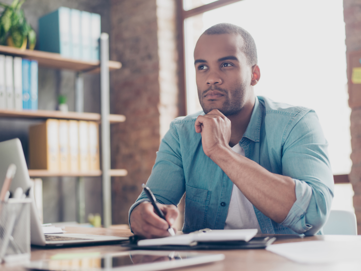 A man in a teal shirt sits at a desk with his hand on his chin, looking thoughtfully into the distance while holding a pen.