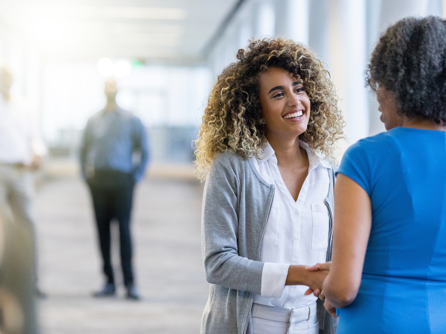 Smiling woman with curly hair shakes hands with another woman in a blue dress in a hallway.