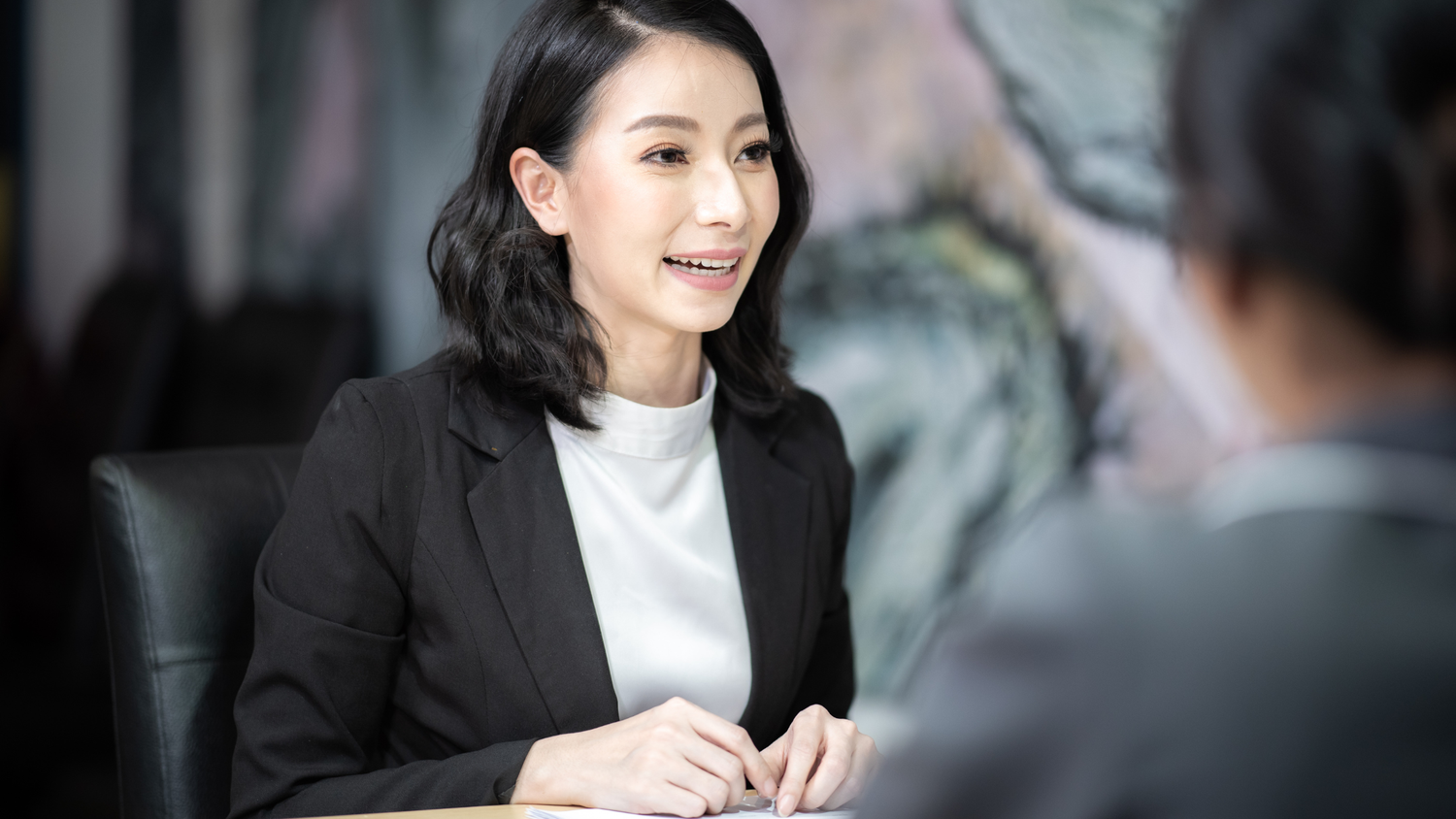 A smiling Asian woman in a black blazer and white shirt talking during a business meeting.