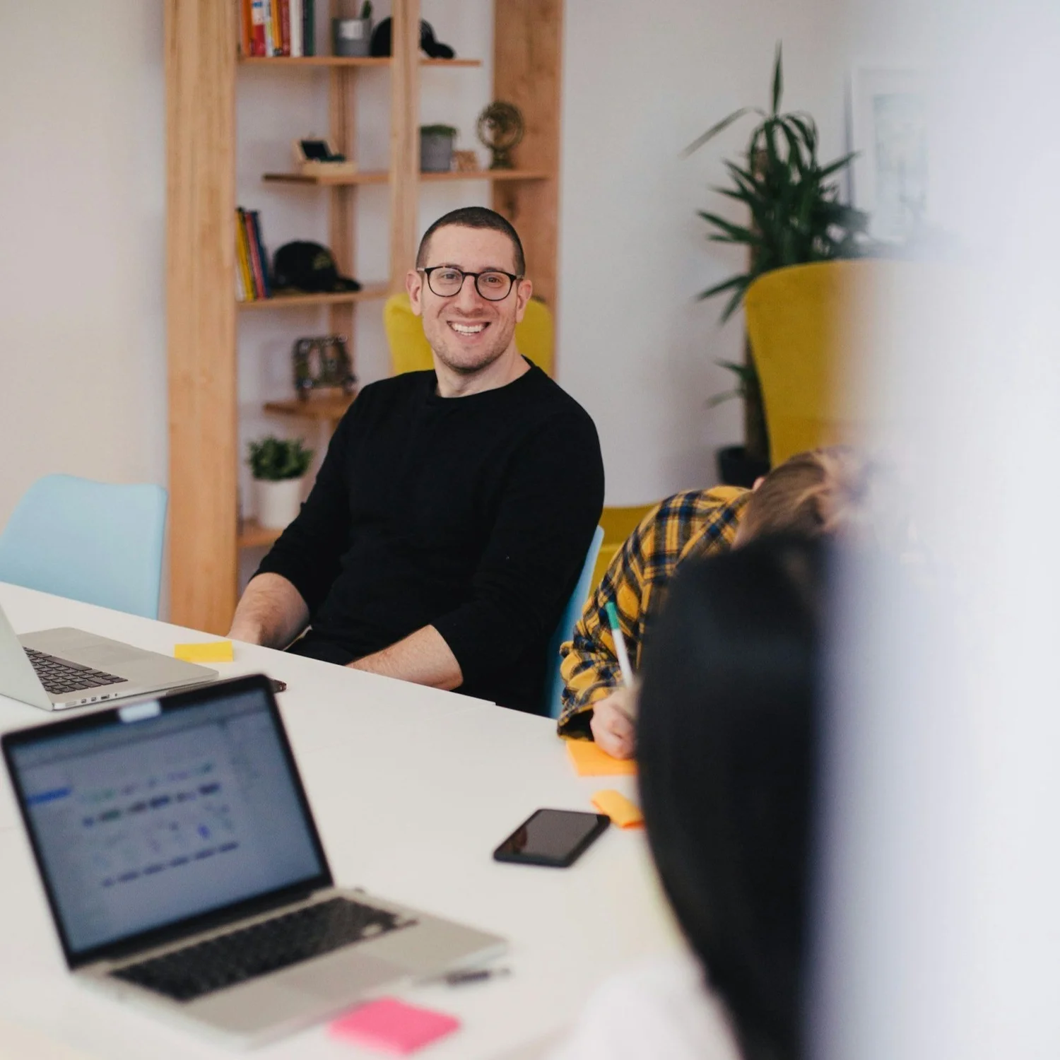 A smiling man in glasses and a black sweater sits at a table with laptops in front of him in an office.