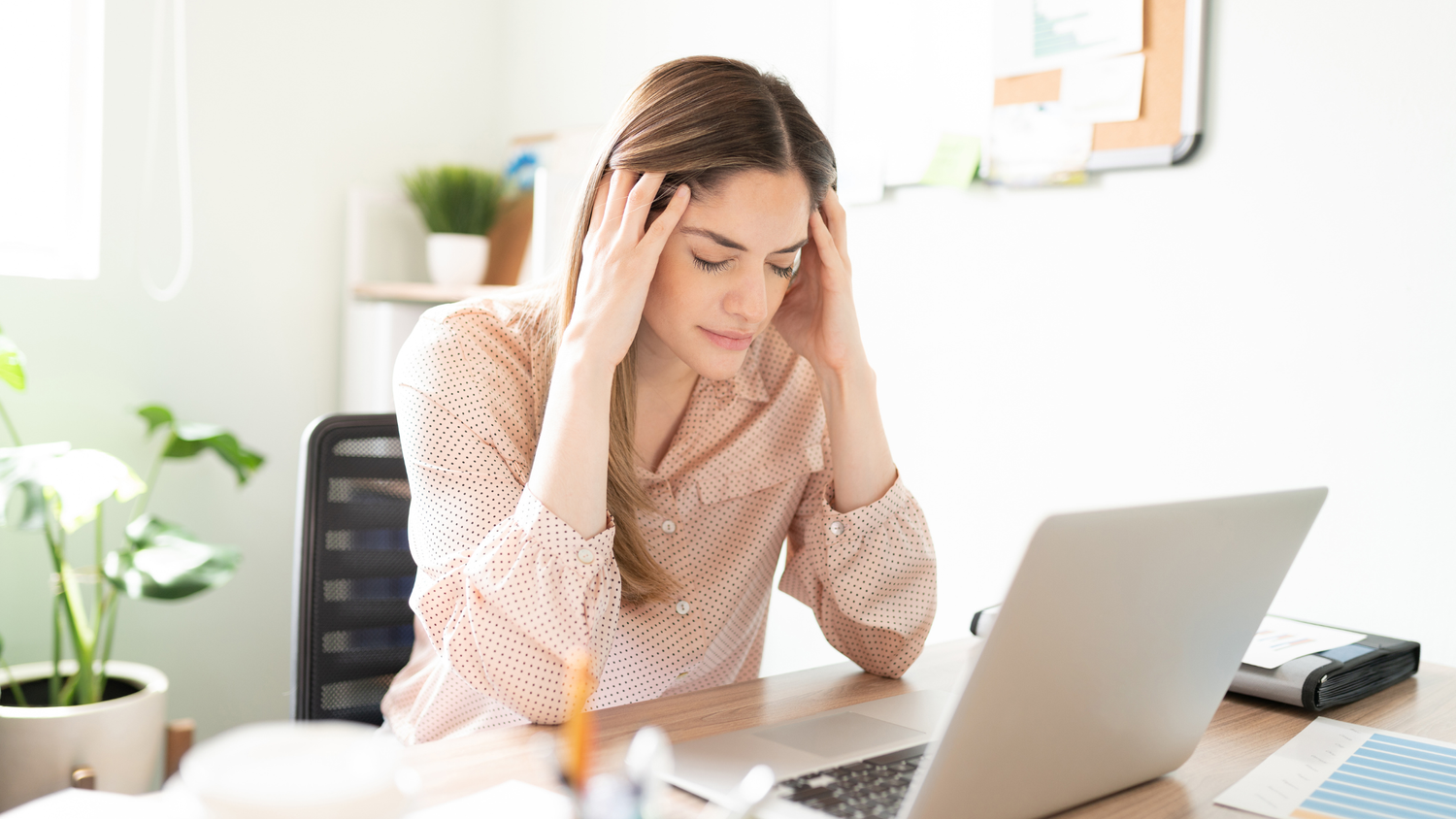 Woman sits at a desk with her eyes closed and hands on her temples, looking stressed while working on a laptop.