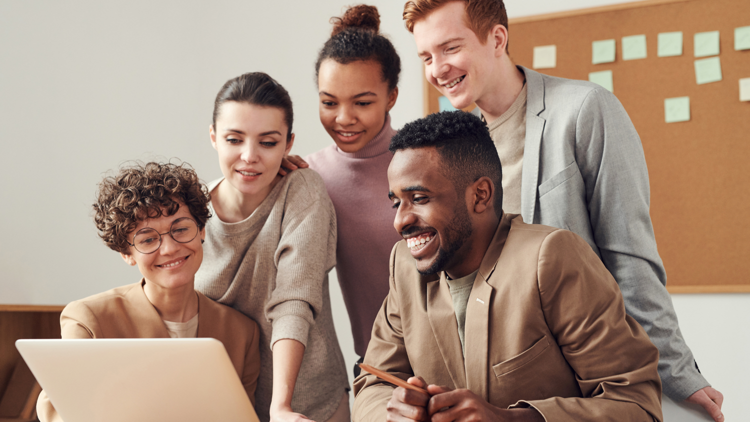 A diverse group of five colleagues collaborate around a laptop, smiling and engaged by the screen.