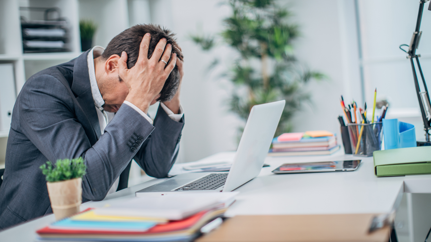 A man in a suit sits at a desk with his head in his hands, looking stressed, a laptop and office supplies nearby.