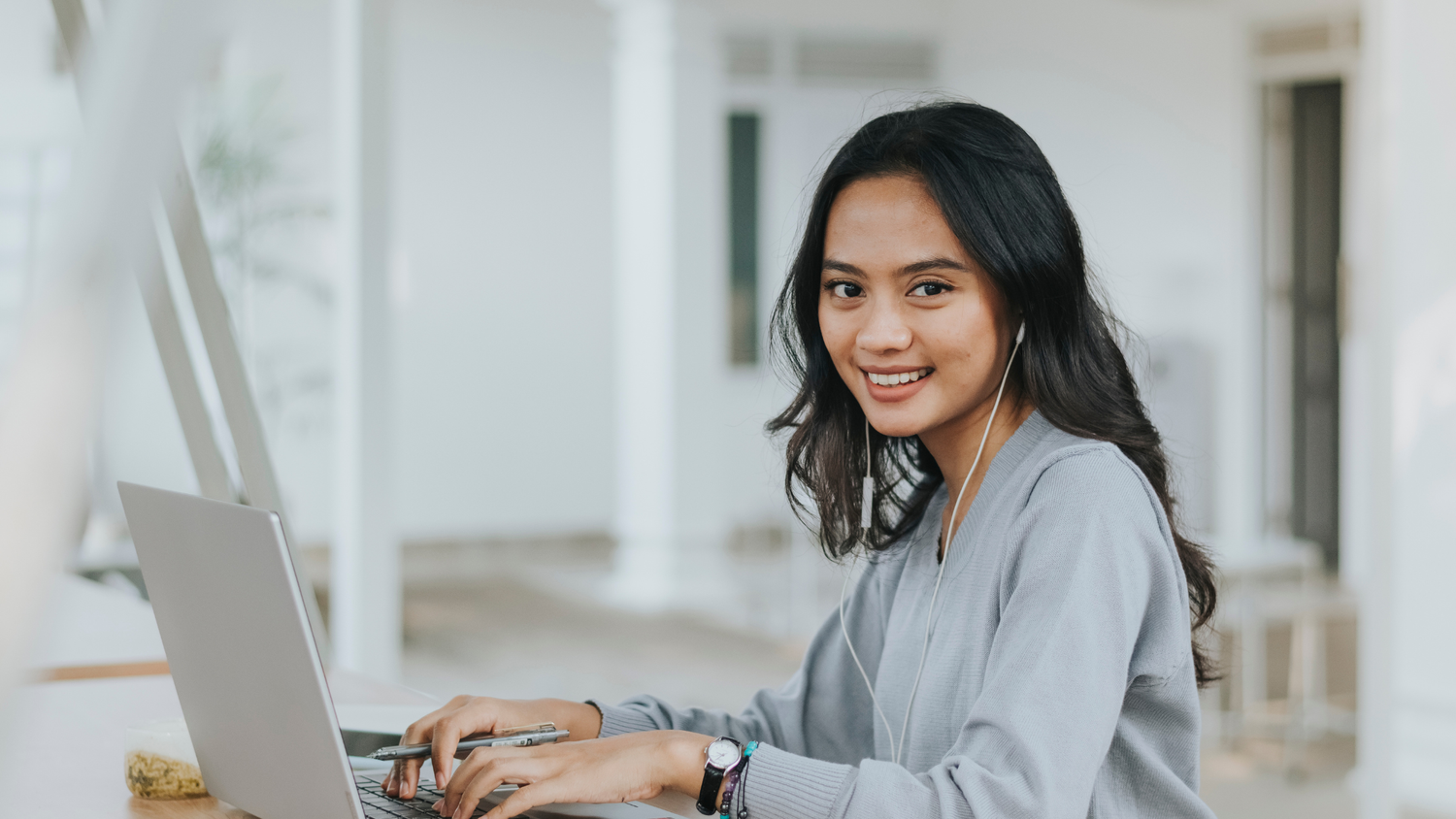 Woman with long dark hair wearing earphones smiles at the camera while typing on a laptop.