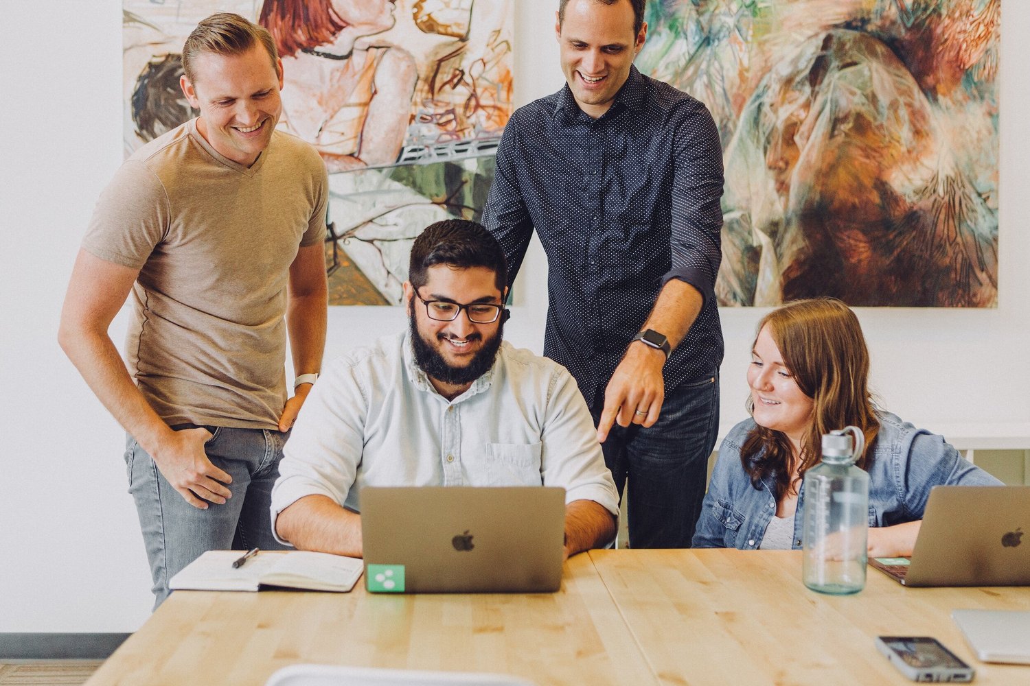 Four smiling colleagues gathered around a table, looking at a laptop.