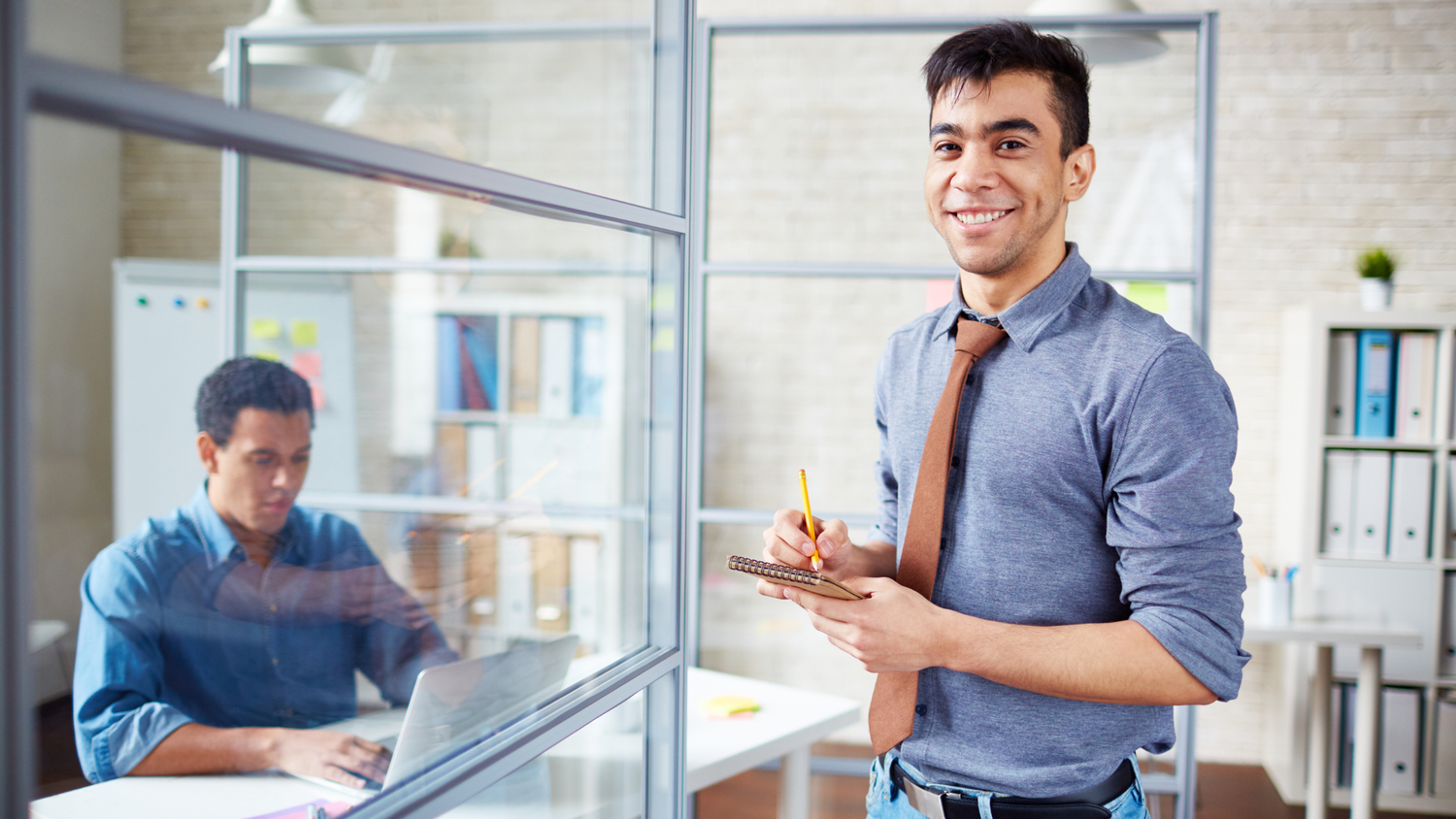 Smiling man in a blue shirt and brown tie holds a notepad and pencil; another man works on a laptop in the background.