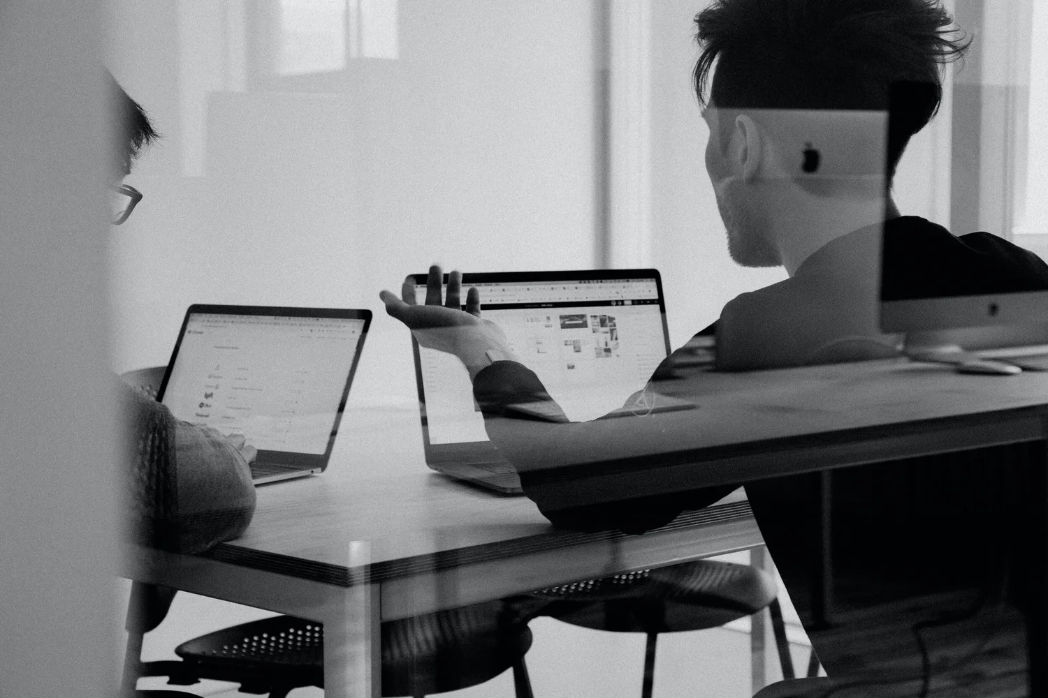 Two people in an office collaborate around laptops, one gesturing to a screen; the image is black and white.
