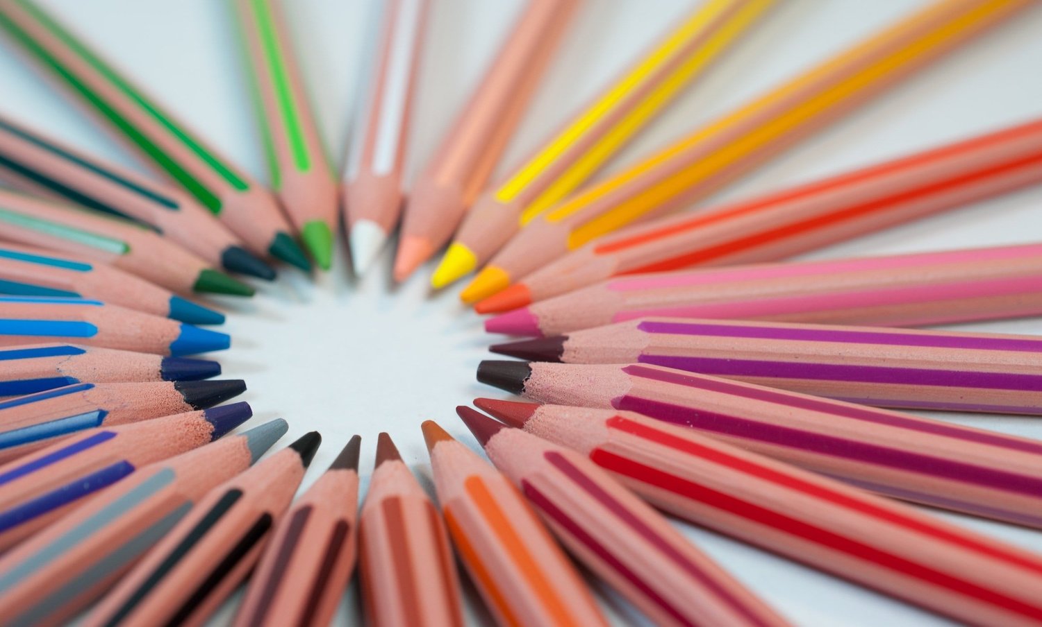Colored pencils arranged in a circle with their tips pointing inward, in shades of green, blue, purple, pink, red, orange, yellow, and peach.