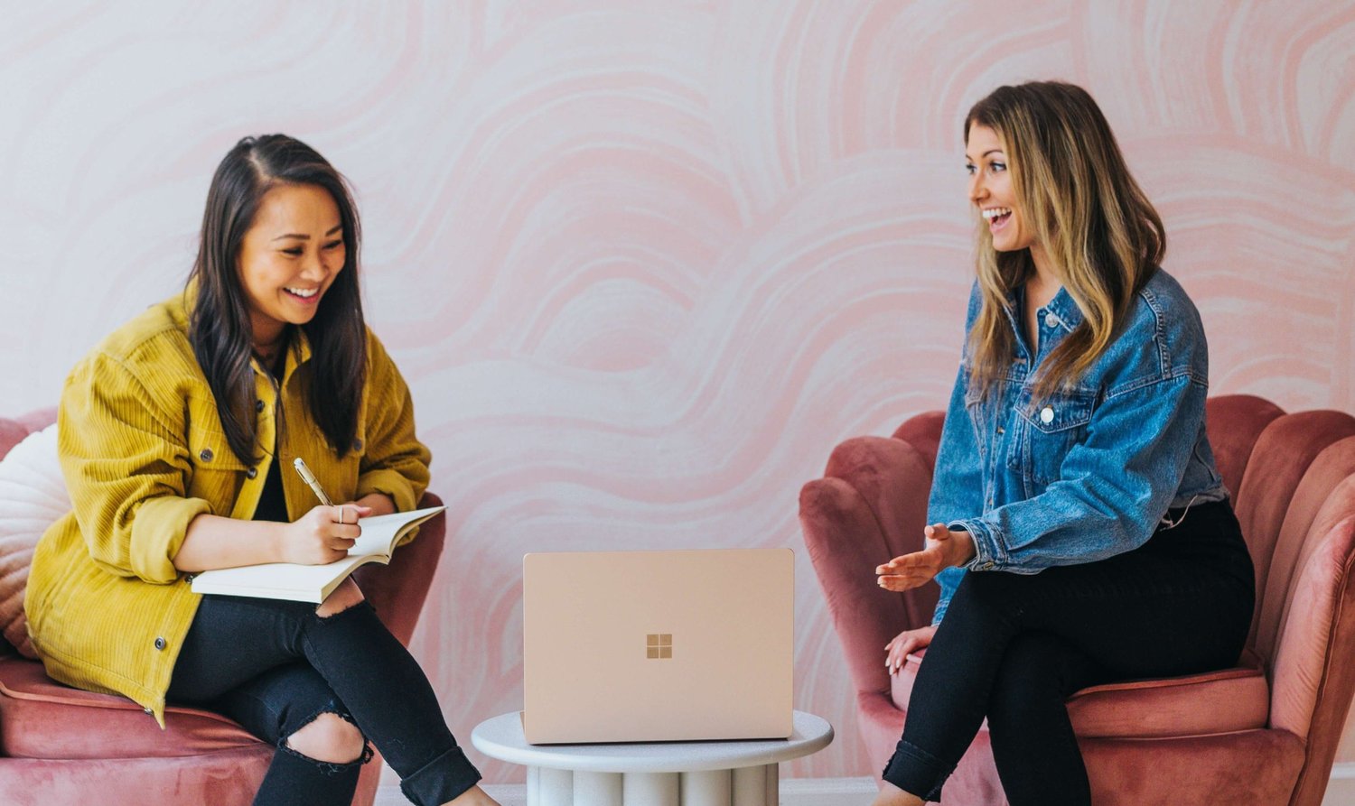 Two women sitting in pink chairs, one journaling and the other gesturing while talking, a laptop on a small table between them.
