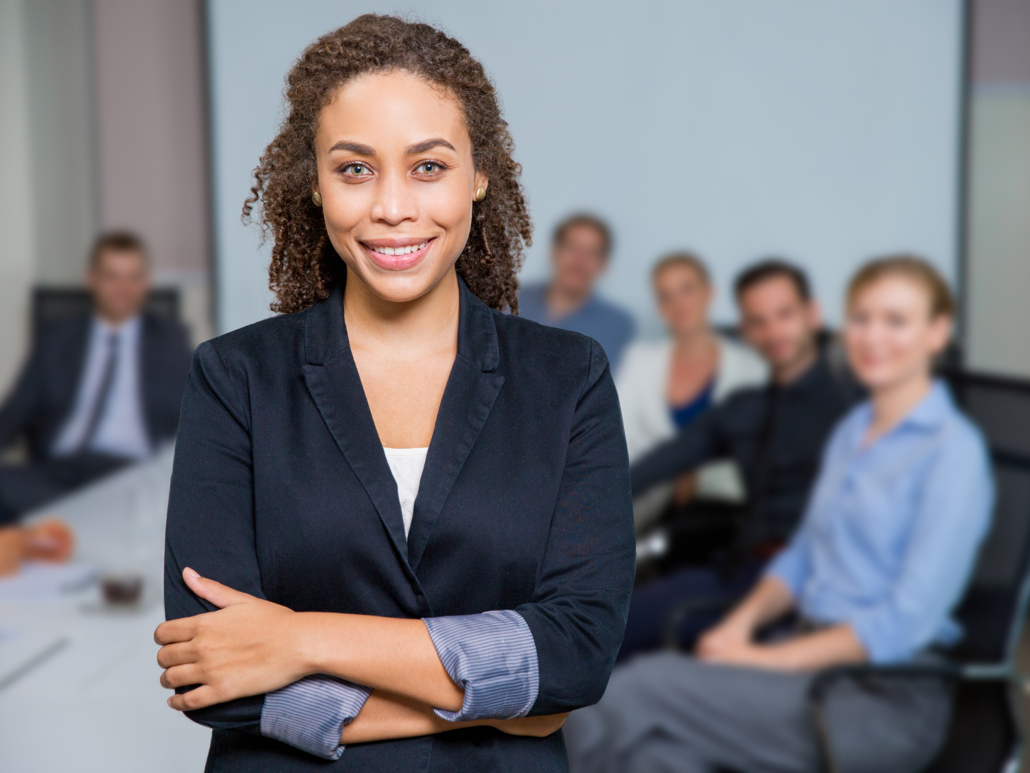 Confident businesswoman with arms crossed in front of a blurred group of people in a meeting room.