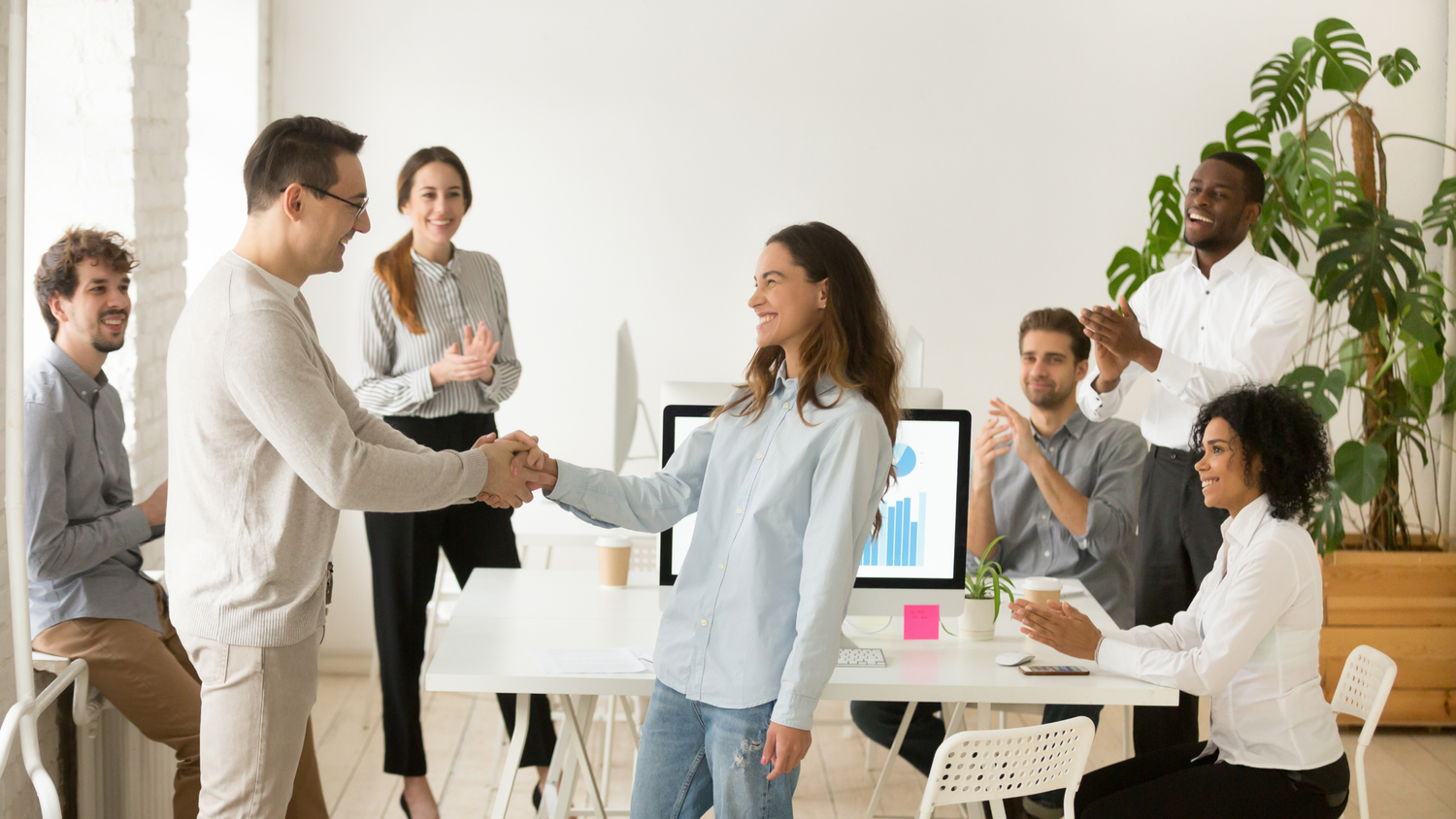 A man and woman shake hands in an office while colleagues clap.