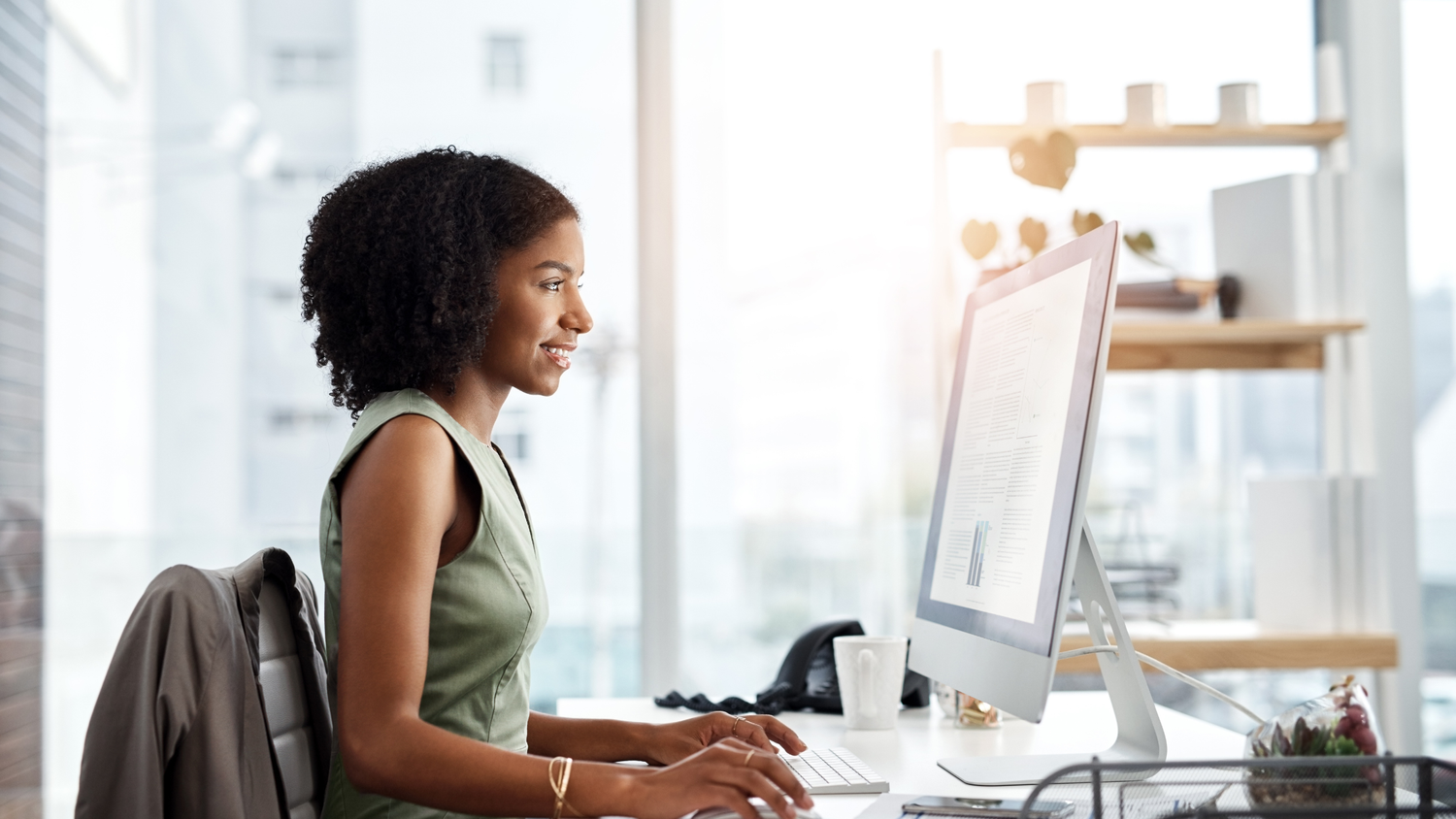 Woman in a green sleeveless top with curly hair smiles while typing on a computer at her desk in a bright office.