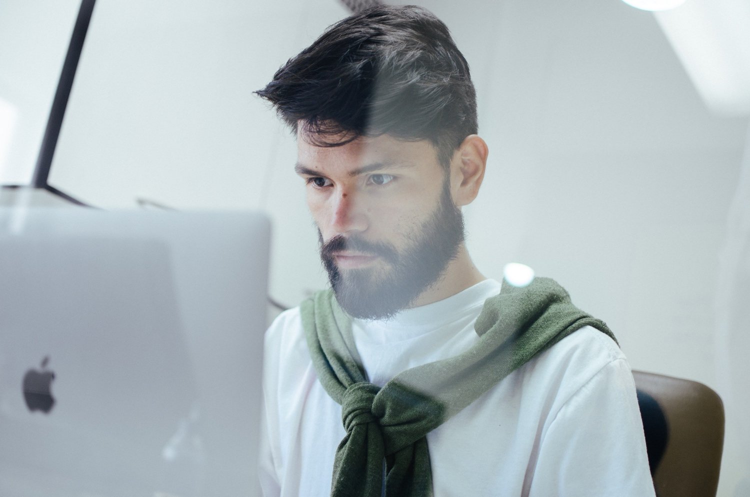 Man with dark hair and a beard focused on a computer screen, wearing a white t-shirt with a green sweater tied around his neck.