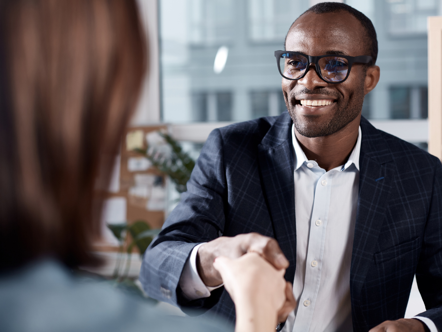 A smiling Black man in a suit and glasses shakes hands with someone across a table.