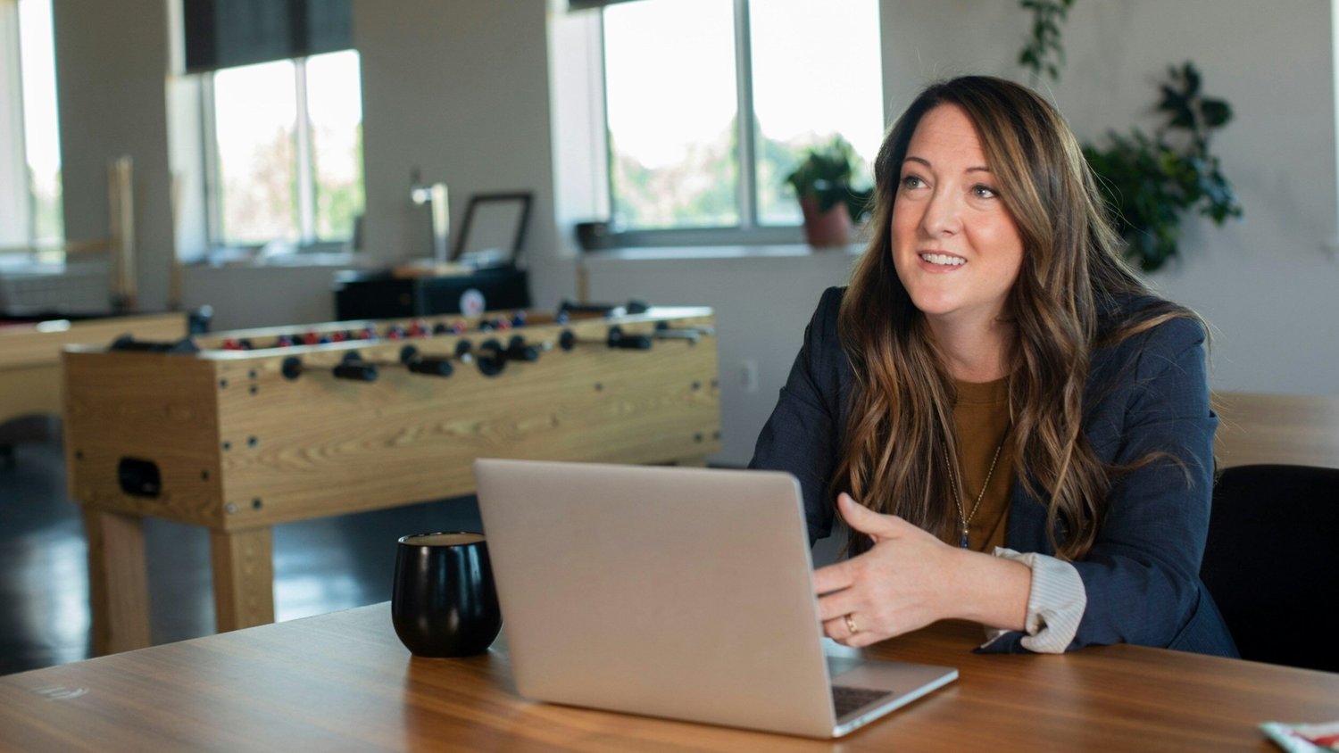 A woman in a navy blazer smiles and gestures while looking away from her laptop, a foosball table in the background.