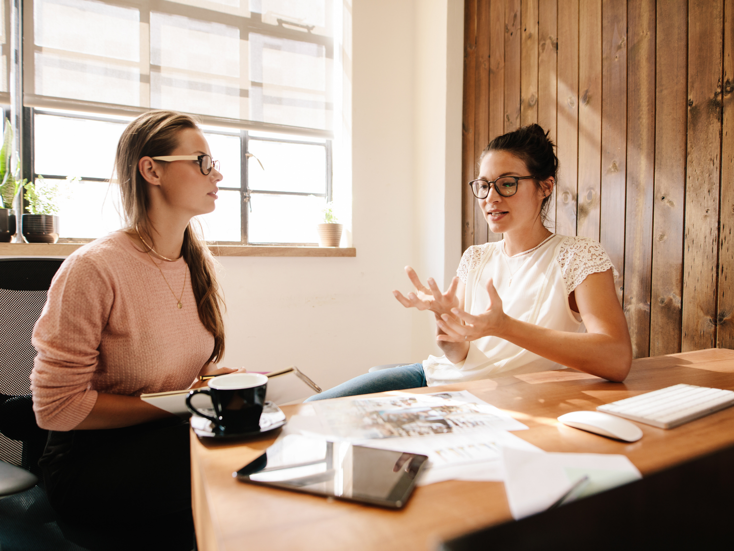 Two women in glasses having a meeting at a wooden desk with papers and a computer.
