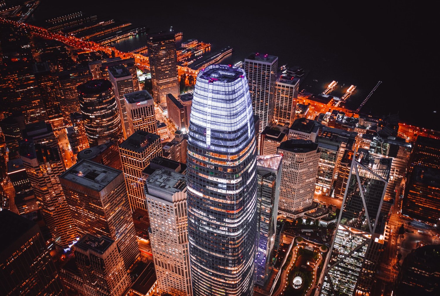 An aerial nighttime view of the Salesforce Tower illuminated in white and blue, surrounded by glowing skyscrapers.
