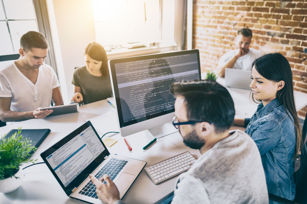 A team of developers reviewing HTML markup on a desktop monitor and a laptop in a bright open-plan office