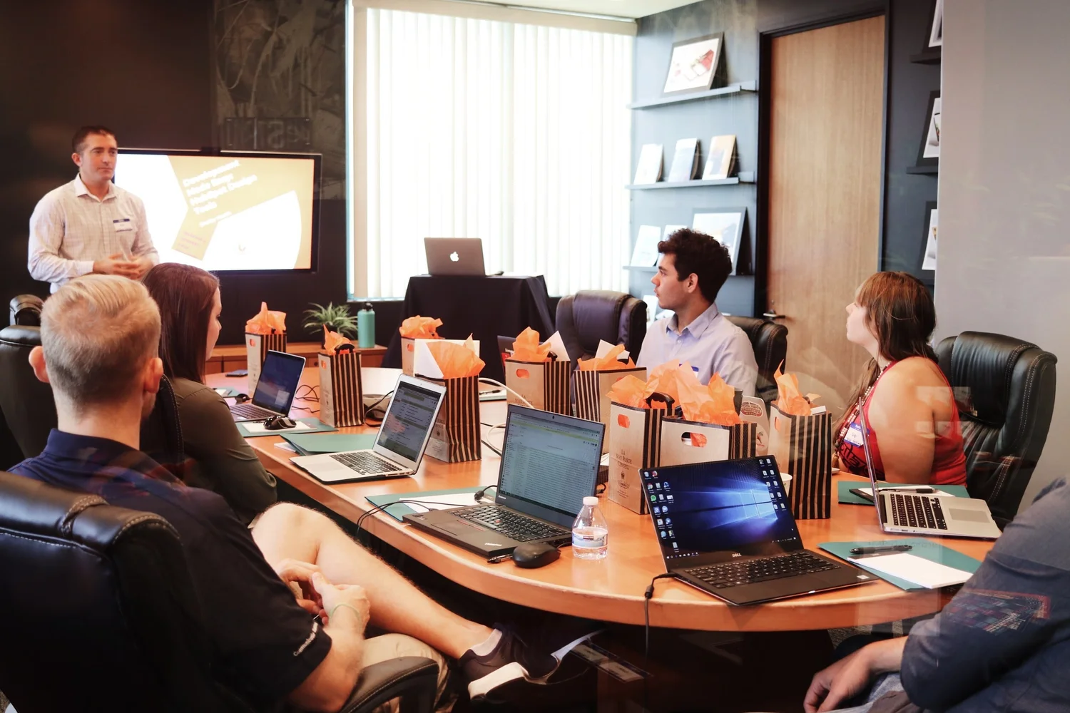 A presenter stands by a screen as a team sits around a conference table with laptops and gift bags.